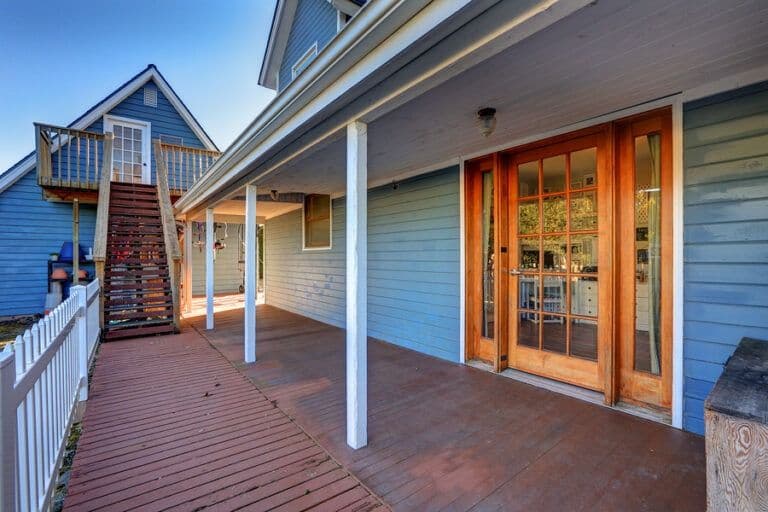 Covered deck with wooden flooring and white support columns, blue house exterior with orange glass doors, white railing, and stairs leading to upper deck and blue porch