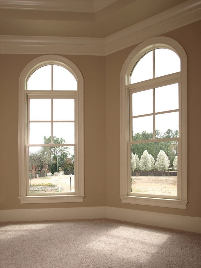 Two tall arched windows with wood frames in a beige room, showing an exterior landscape with evergreen trees and open grounds
