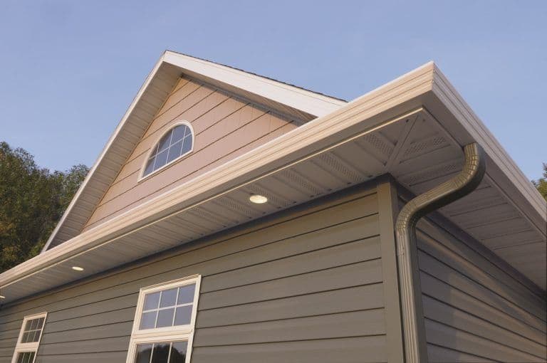 House exterior detail showing peaked roofline with circular window, horizontal siding, rectangular windows, and curved gutter system against clear blue sky