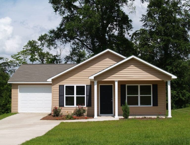 Single-story residential home with tan siding, white trim, and gray roof, featuring a front porch, white garage door, and black shutters surrounded by mature trees and green lawn
