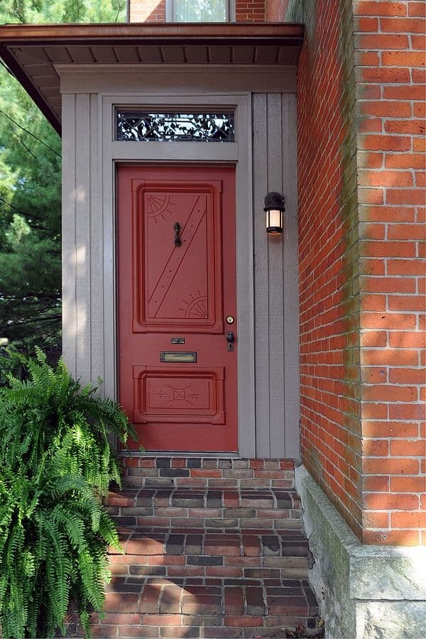 Red front door with decorative panels and transom window, set in gray frame on brick house with wall sconce light and fern planter
