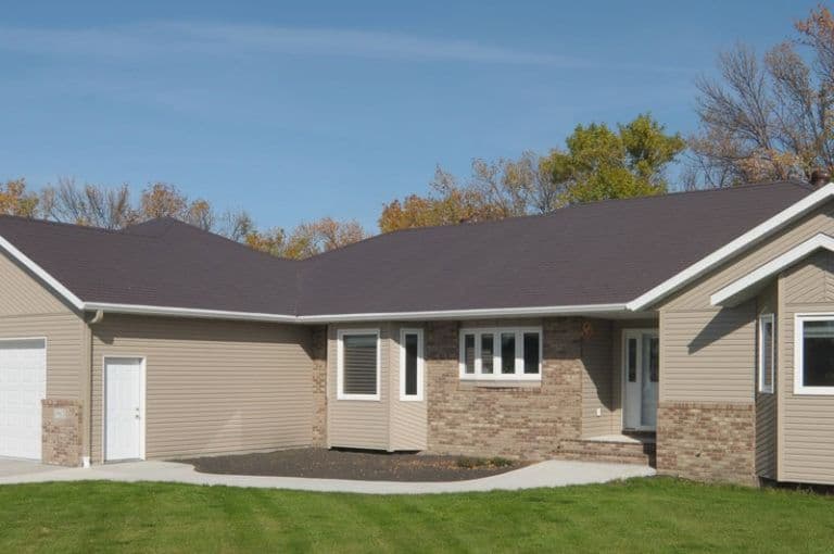 Modern single-story residential home with tan siding, brick accents, dark roof, attached garage, and front entrance surrounded by autumn trees
