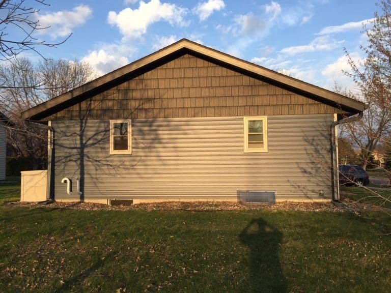 Single-story residential building with tan vinyl siding, brown shingle roof, two windows, and bare trees in yard