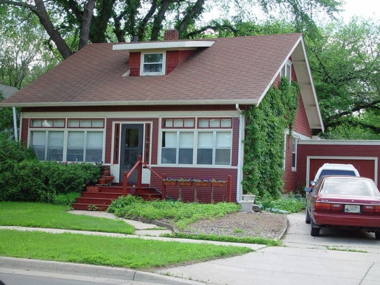 Red brick residential home with large front windows, front porch steps, ivy-covered side wall, mature tree overhead, and red sedan parked in attached garage driveway
