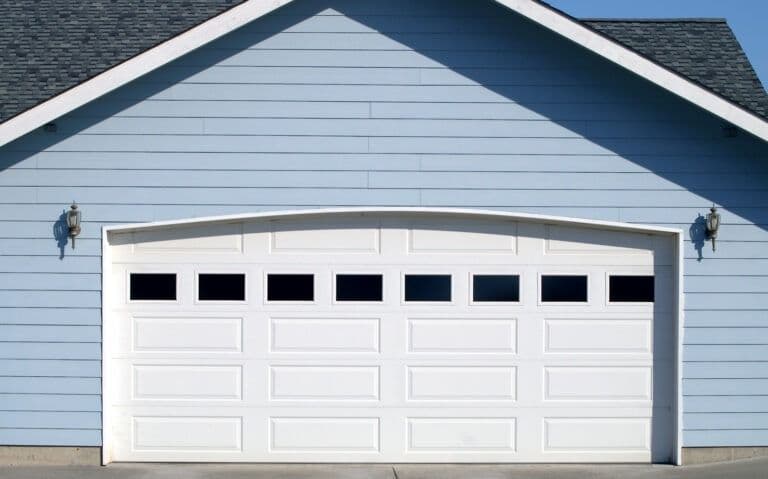 White two-car garage door with eight panels and decorative windows on a light blue house with gray trim and wall-mounted lights light blue