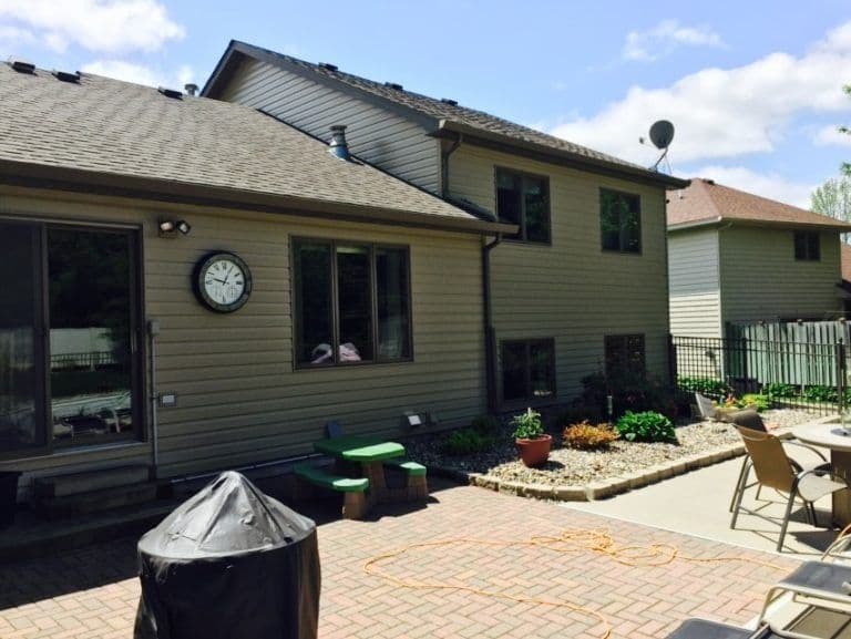 Two-story beige house with gray roof, patio with brick pavers, outdoor furniture, and planters, satellite dish visible