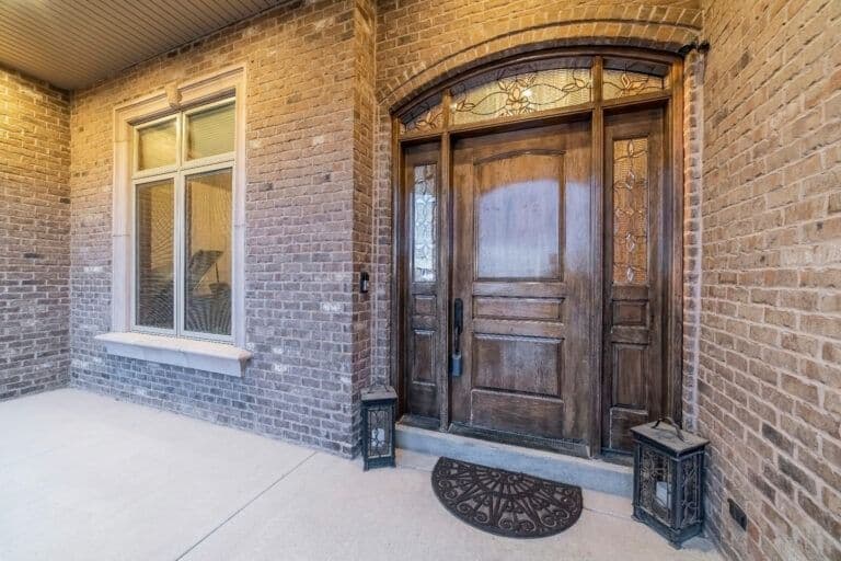 Brick entryway with wooden front door, sidelights, transom window, and decorative welcome mat flanked by planters
