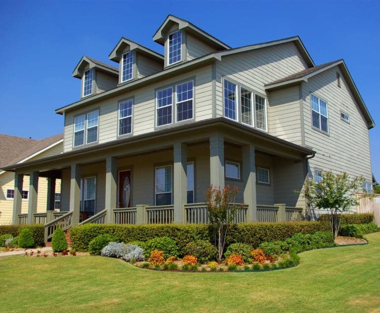 Two-story cream-colored house with three dormer windows, front porch with columns, and landscaped yard featuring shrubs and flowering plants under blue sky