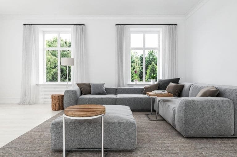 Bright minimalist living room with gray sectional sofa, wooden side tables, and two large windows with sheer curtains overlooking greenery