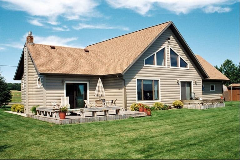 Single-story residential home with tan log siding, peaked roof, front porch with deck railing, large windows, and manicured lawn under clear blue sky
