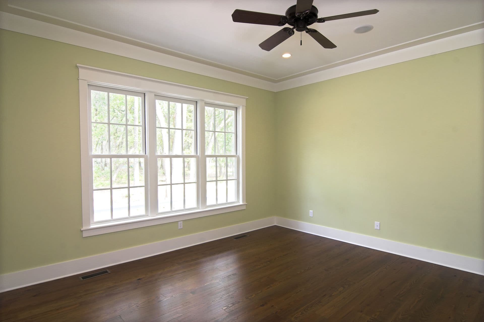 Empty bedroom with pale green walls, dark wood flooring, ceiling fan, and large triple-pane windows overlooking trees