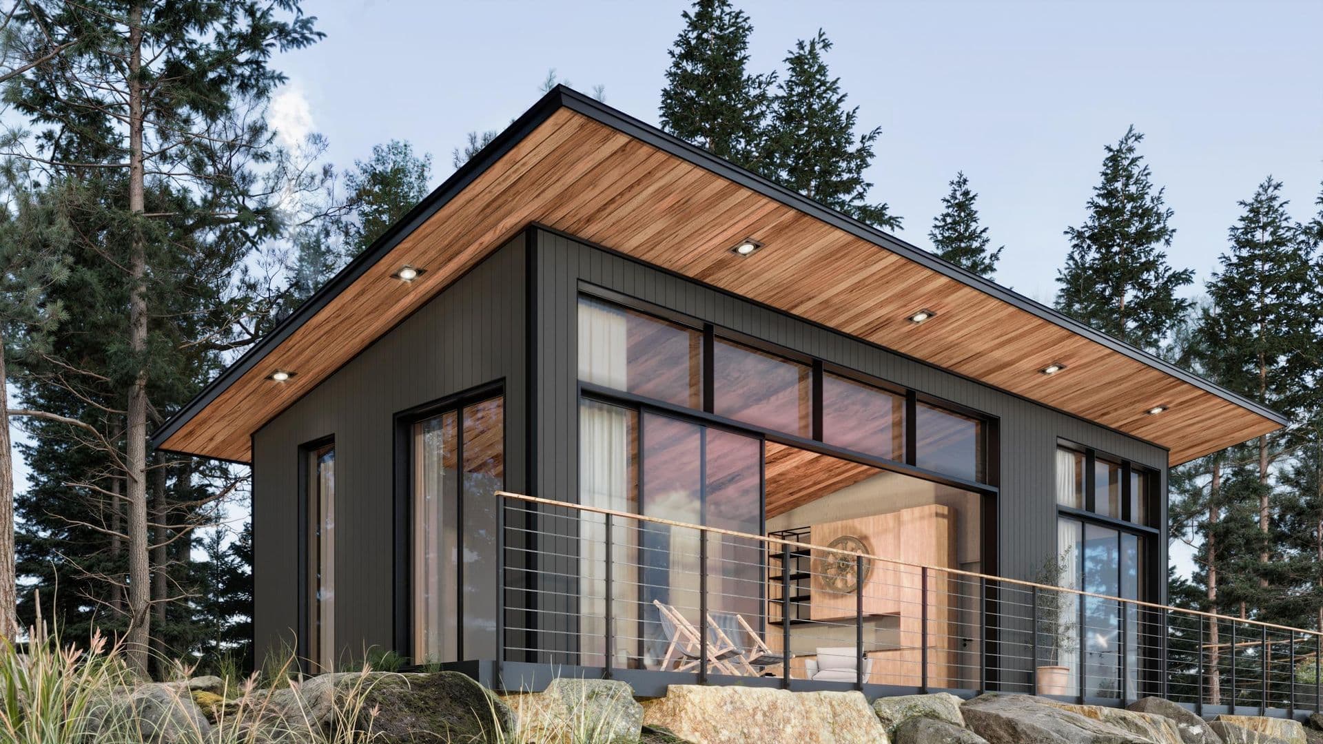 Modern cabin with black metal frame, wood ceiling overhang, and expansive glass wall overlooking forest surrounded by tall evergreen trees and boulders