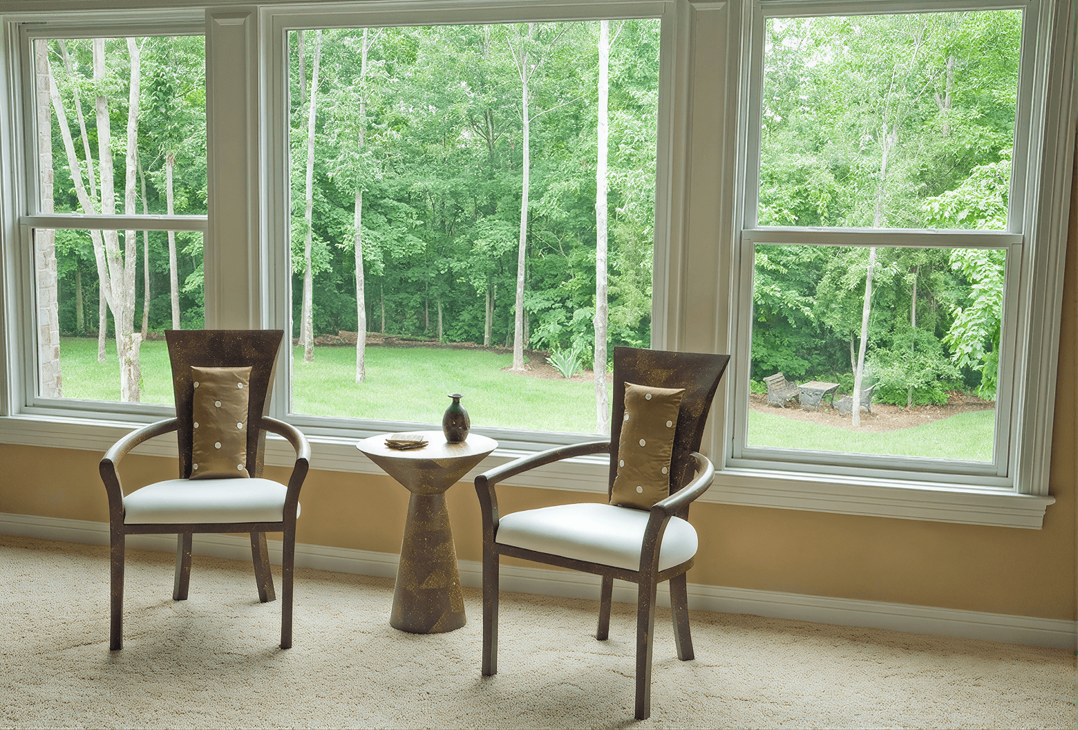 Seating area with two upholstered armchairs flanking a round pedestal table facing large windows with views of a forested landscape