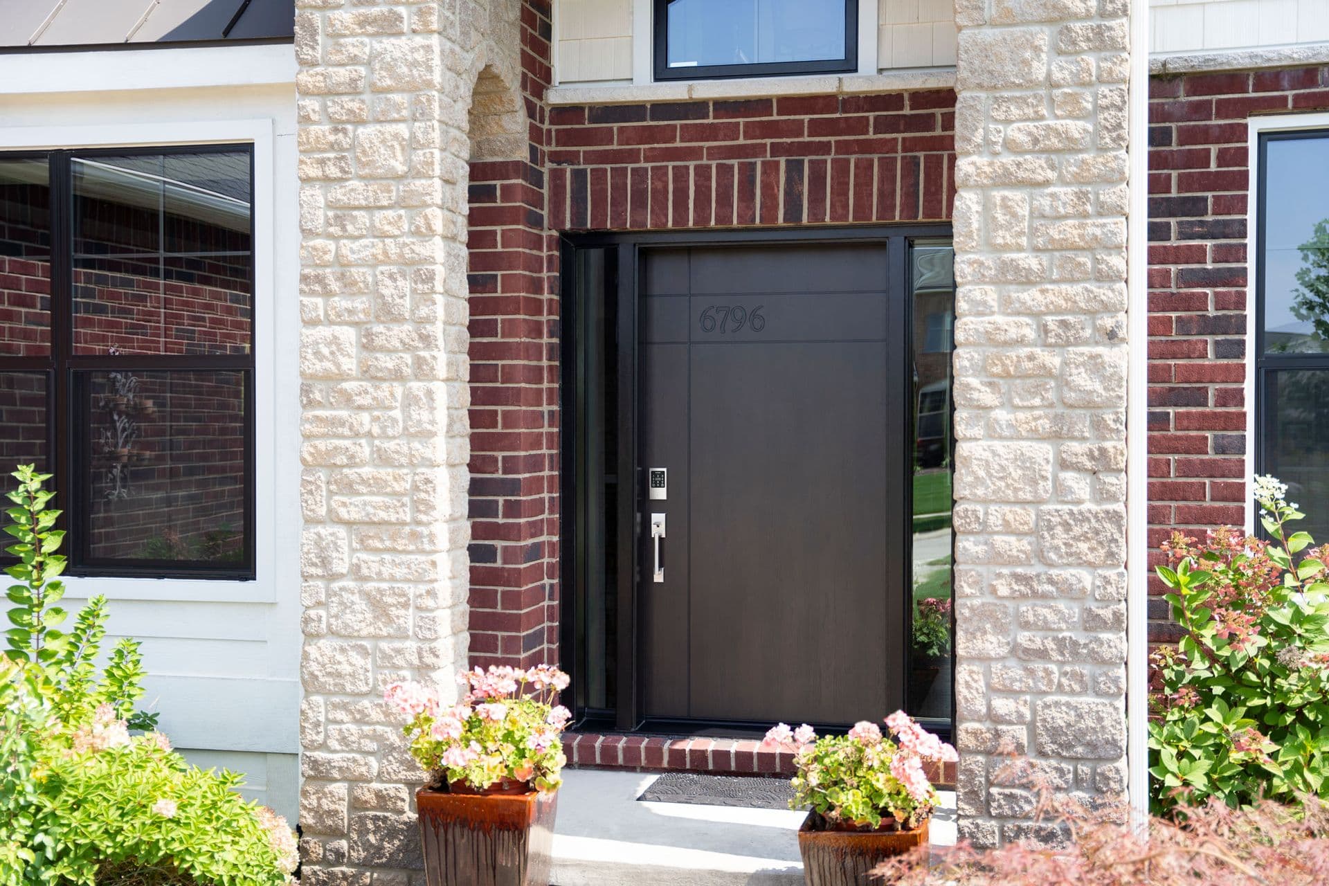 Residential home entrance with black front door marked 6796, flanked by cream and red brick columns, potted pink flowers, and contemporary windows