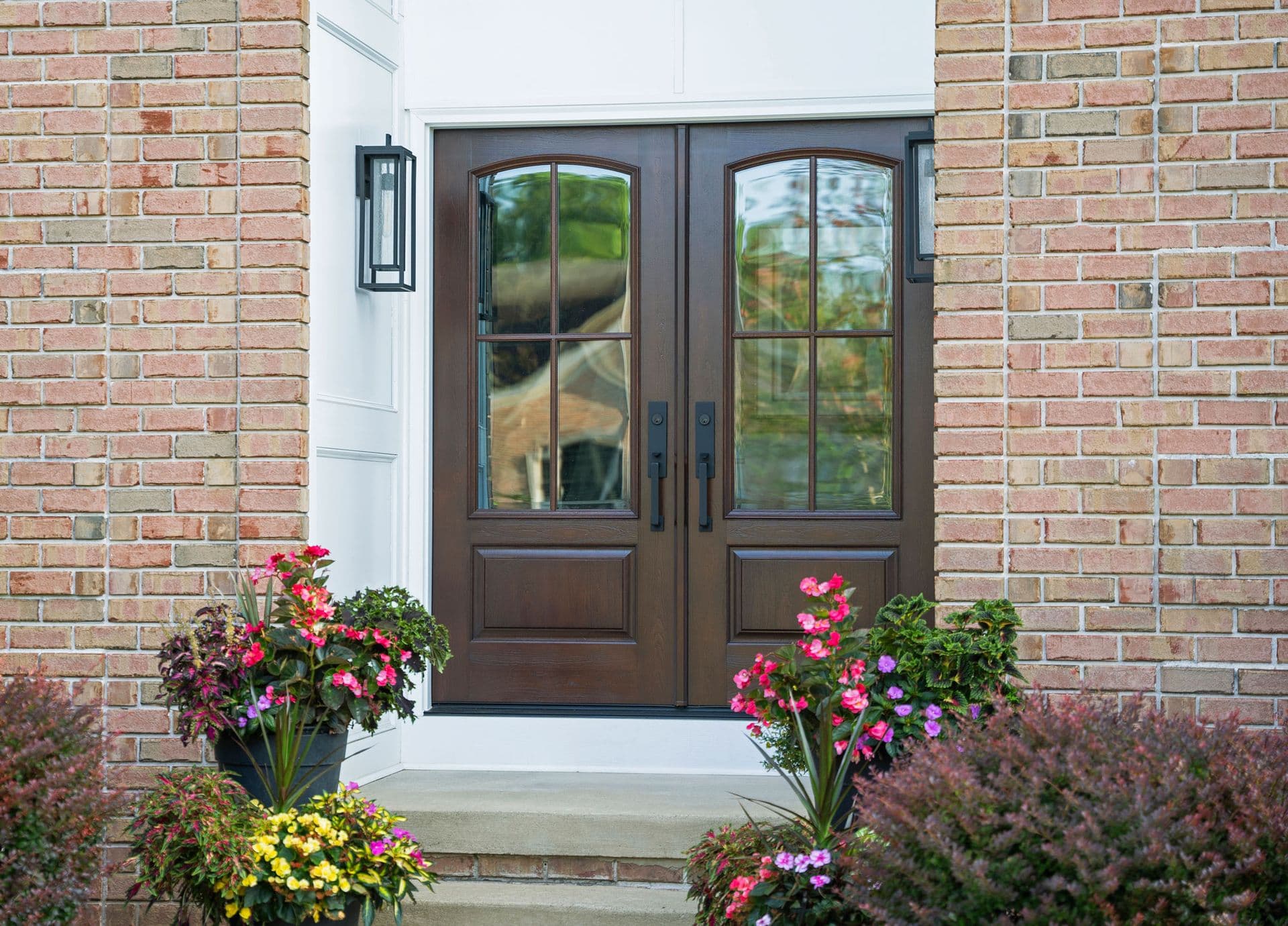 Dark brown double entry doors with glass panes on a brick home, flanked by colorful flowering planters in pink, purple, and yellow