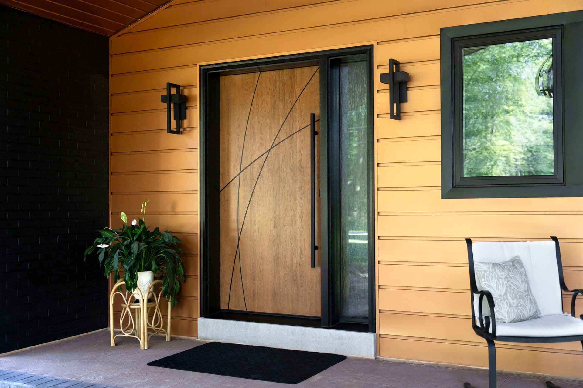 Modern home entrance with yellow horizontal siding, wooden front door with black frame, decorative wall sconces, potted plant, and patio seating