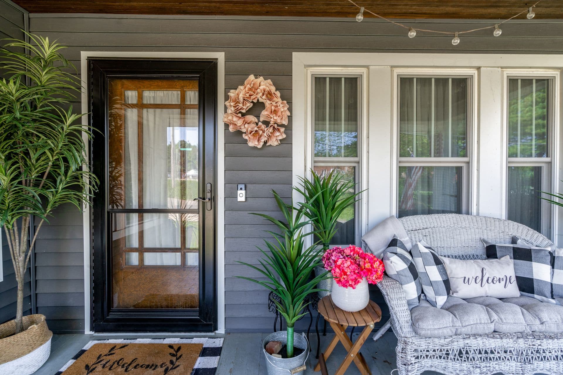 Covered porch with black entry door, wicker seating, potted plants with pink flowers, peach wreath, and welcome pillow on gray cushioned furniture