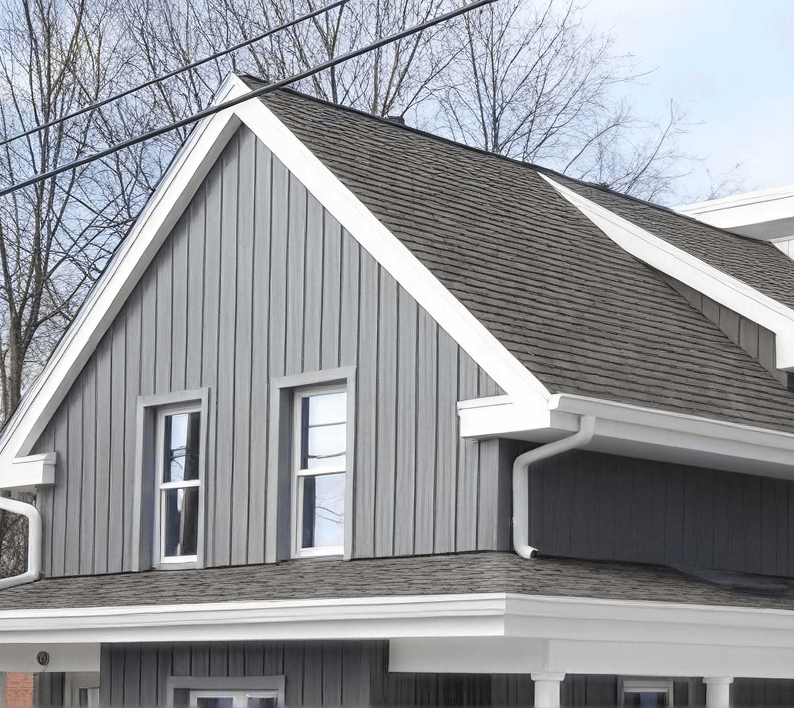 Modern farmhouse exterior with gray vertical siding, white trim, two upper windows, dark shingle roof, attached garage, and bare trees in background